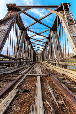 The bridge is old and rusted, with a wooden structure and a metal rail. The sky is blue and clear, and the bridge is surrounded by treesの写真素材