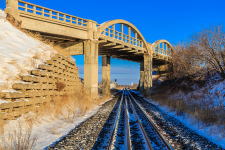 A train tracks are shown in the snow with a bridge in the background. The bridge is a large arch and...
