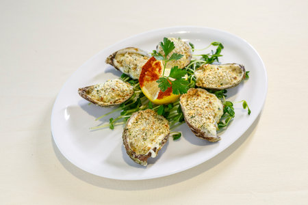 A plate of oysters with a lemon wedge and parsley on top. The plate is white and the oysters are...