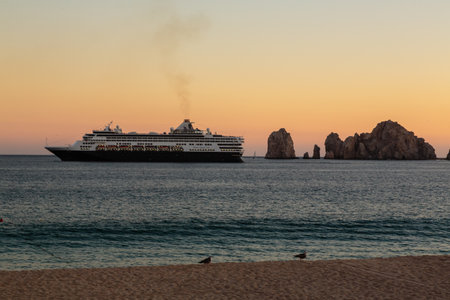 A large cruise ship is sailing in the ocean near a rocky shore. The sky is a beautiful mix of orange...