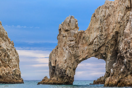 A rocky cliff with a large archway in the middle. The archway is surrounded by rocks and the water is calmの写真素材
