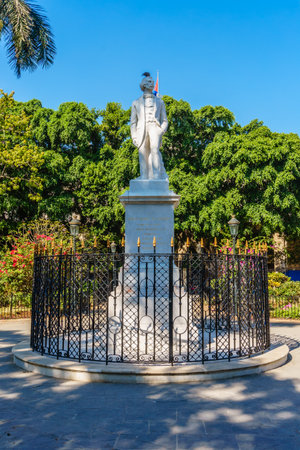 A statue of a man stands in front of a fence. The statue is white and the fence is black. The statue is surrounded by a green area with treesの写真素材