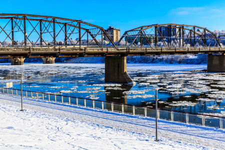 A bridge spans a river with a snowy bank on either side. The bridge is surrounded by a fence and a sidewalk. The sky is clear and the water is frozenの写真素材
