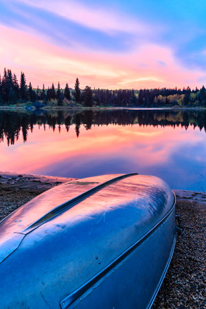 A canoe is sitting on the shore of a lake at sunset. The sky is a beautiful mix of pink and orange hues, creating a serene and peaceful atmosphere. The reflection of the treesの写真素材