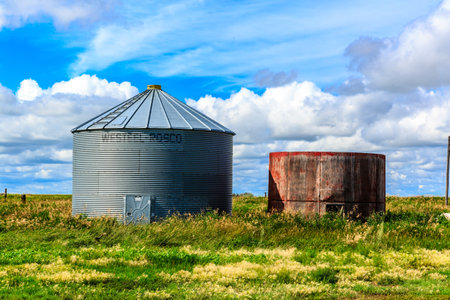 A large metal silo sits in a field next to a smaller, rusty metal silo. The sky is cloudy, but the sun is still shining through the clouds. The scene is peaceful and quietの写真素材