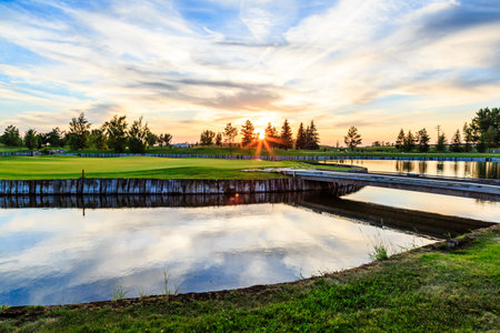 A golf course with a bridge over a pond and a sunset in the background. The water is calm and the sky is filled with cloudsの写真素材