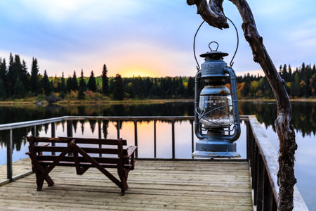 A lantern hangs from a tree branch over a dock by a lake. The lantern is lit, and the water is calm....