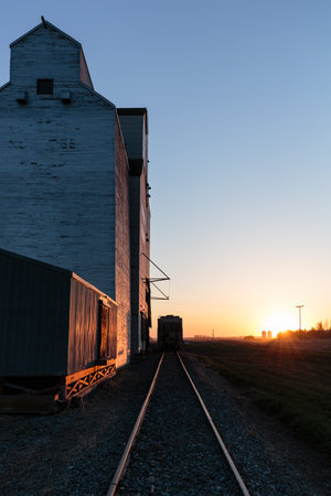 A train is traveling down a track next to a grain silo. The sun is setting in the background, casting a warm glow over the sceneの写真素材