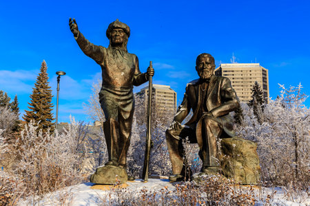 Two statues of men sitting on a rock in a snowy park. One of the statues is holding a walking stickの写真素材