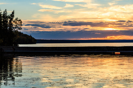 The sky is a beautiful orange color as the sun sets over the water. The water is calm and still, reflecting the sky and the trees in the background. The scene is peaceful and sereneの写真素材