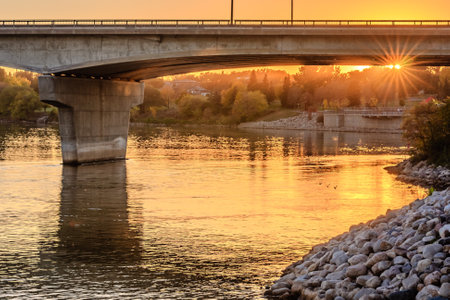 A bridge spans a river with a sunset in the background. The sun is shining brightly on the water, creating a beautiful and serene atmosphereの写真素材