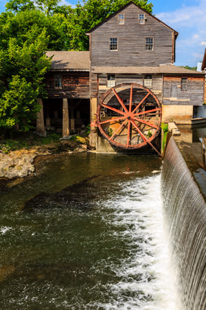 A large water wheel is in the foreground of a river. The water wheel is surrounded by a small...