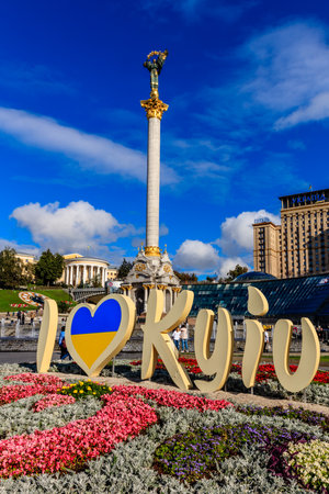 A large, white, and blue sign with the word "I love Kyiv" written on it. The sign is placed in front of a beautiful flower garden, with a tall, white, and blue column in the backgroundの写真素材