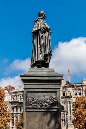 A statue of a man stands in front of a building. The statue is made of bronze and is very detailed. The sky is blue and there are some clouds in the backgroundの写真素材