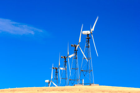 A row of wind turbines are on a hill, with the sun shining brightly in the background. The turbines are tall and spread out, with some of them being closer to the camera than othersの写真素材