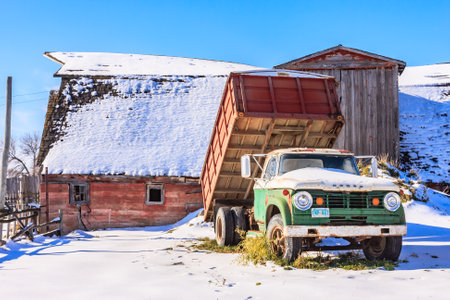 An old green truck is parked in the snow next to a red barn. The truck is covered in snow and he is abandoned. Concept of nostalgia and a feeling of being in a rural, wintery settingの写真素材