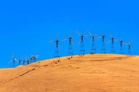 A row of windmills are on a hillside. The windmills are tall and spread out. The sky is blue and...