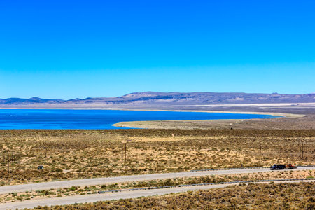 A car is driving down a road next to a body of water. The sky is clear and the sun is shining brightlyの写真素材