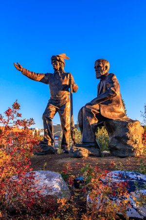 Two statues of men, one of them holding a walking stick, are sitting on a rock. The other statue is standing. The sky is blue and the sun is settingの写真素材
