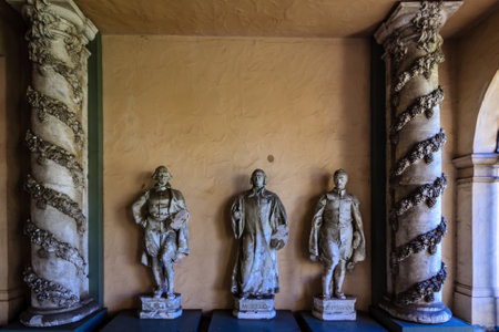 Three statues of men are standing in front of two pillars. The statues are of different sizes and are made of stone. The pillars are decorated with vines and leavesの写真素材
