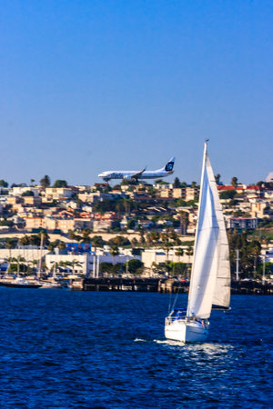 A sailboat is sailing in the ocean with a plane flying in the background. The scene is peaceful and serene, with the water and sky blending together to create a calming atmosphereの写真素材