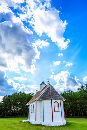 A small white church with a red roof sits in a field. The sky is cloudy, but the sun is shining through the clouds, creating a warm and peaceful atmosphereの写真素材