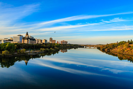 A city skyline is reflected in the water of a river. The sky is clear and blue, and the water is calmの写真素材