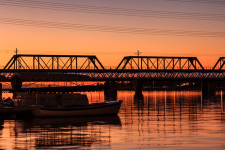 A bridge spans a river with a boat in the water. The sky is orange and the water is calm