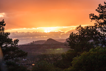 The sun is setting over a mountain range, casting a warm glow over the landscape. The sky is filled with clouds, creating a serene and peaceful atmosphere. The trees in the foreground are lushの写真素材