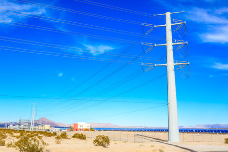 A tall power pole stands in the desert, with a clear blue sky above. The pole is surrounded by a fence, and there is a small building in the distance. Concept of isolation and emptinessの写真素材