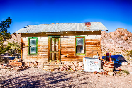 A small, old house with a tin roof sits on a dirt road. The house is surrounded by a few potted plants and a refrigerator. Scene is one of solitude and simplicityの写真素材