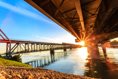 A bridge spans a river with a train passing underneath. The sun is shining brightly, creating a warm and inviting atmosphereの写真素材