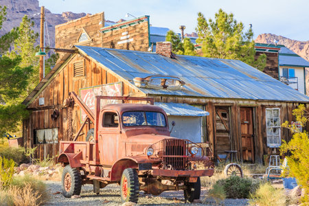 An old truck is parked in front of a rustic building. The truck is a red pickup truck with a large bed. The building is a small house with a tin roof. The scene is set in a desertの写真素材