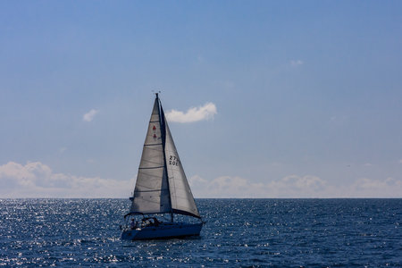 A sailboat is sailing in the ocean with a clear blue sky above. The sailboat is white and has the...