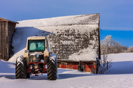 A tractor is parked in front of a red barn. The tractor is covered in snow and the barn is also covered in snow. The scene is quiet and peaceful, with the snow covering the ground and the barnの写真素材