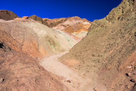 A rocky, hilly landscape with a path running through it. The colors of the rocks are brown, green, and yellow. The sky is blue and clearの写真素材