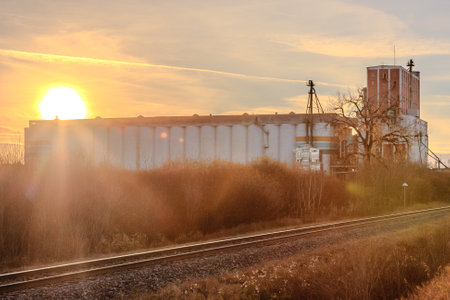 A train tracks run through a field with a large building in the background. The sun is setting, casting a warm glow over the sceneの写真素材