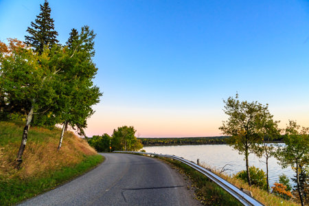 A road with trees on the side and a lake in the background. The sky is blue and the sun is settingの写真素材