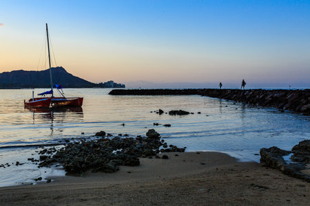 A sailboat is floating in the ocean near a rocky shore. The sky is a beautiful mix of orange and blue hues, creating a serene and peaceful atmosphereの写真素材