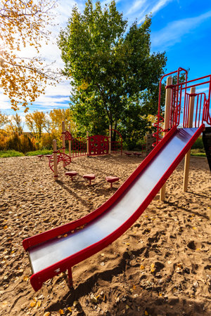 A red and silver playground slide is in a park. The slide is on a sandy surface. The sky is blue and the sun is shiningの写真素材