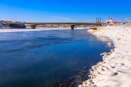 A bridge spans a river with a snowy bank on the other side. The water is frozen and the sky is clearの写真素材