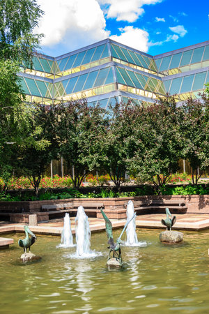 A fountain with four water spouts and a large building in the background. The fountain is surrounded by trees and benchesの写真素材