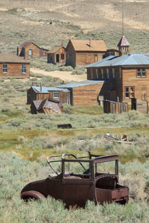 A rusted old car is sitting in a field next to a row of old houses. The scene is quiet and peaceful,...