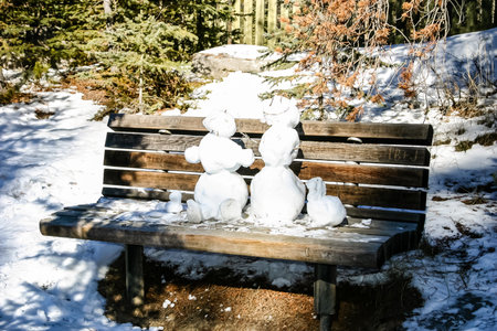 Two snowmen are sitting on a bench in the snow. The bench is wooden and has a rustic feel. The snowmen are small and appear to be made of snow. The scene is peaceful and sereneの写真素材
