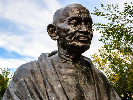A statue of Mahatma Gandhi is sitting in a park. The statue is made of bronze and is very detailed. The sky is blue and there are some clouds in the backgroundの写真素材