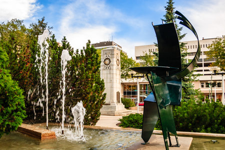 A statue of a man stands in front of a fountain in a park. The fountain is surrounded by trees and has a clock on it. The statue is made of metal and is placed in the middle of the fountainの写真素材