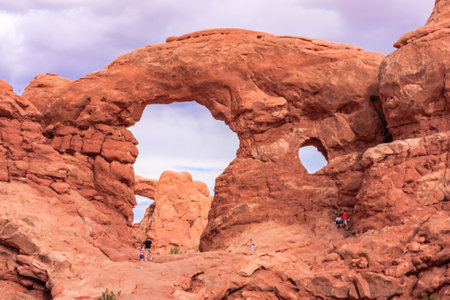 A group of people are walking through a canyon with a large archway in the middle. The archway is surrounded by rocks and the sky is cloudyの写真素材
