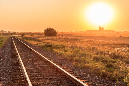 The sun is setting over a train track, casting a warm glow over the landscape. The train tracks are empty, and the sky is a beautiful mix of orange and pink hues. Concept of calm and tranquilityの写真素材