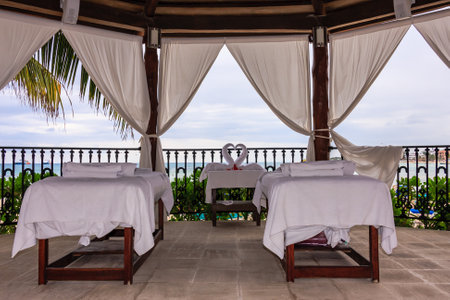 A couple of massage tables are set up on a balcony overlooking the ocean. The tables are covered with white sheets and there are two towels hanging nearby. Concept of relaxation and tranquilityの写真素材