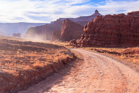 A dirt road winds through a desert landscape with a mountain in the background. The road is dusty and the sky is cloudyの写真素材